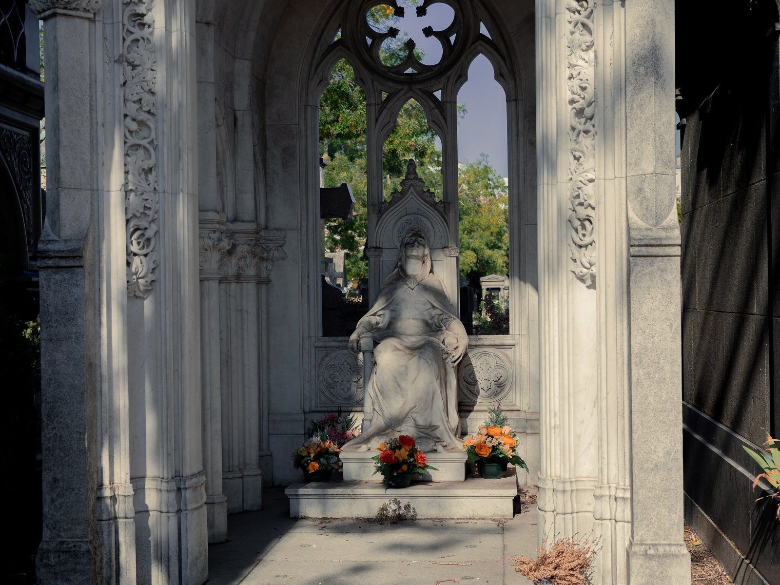 A seated veiled figure in white marble beneath a carved Gothic canopy, with flowers at its feet