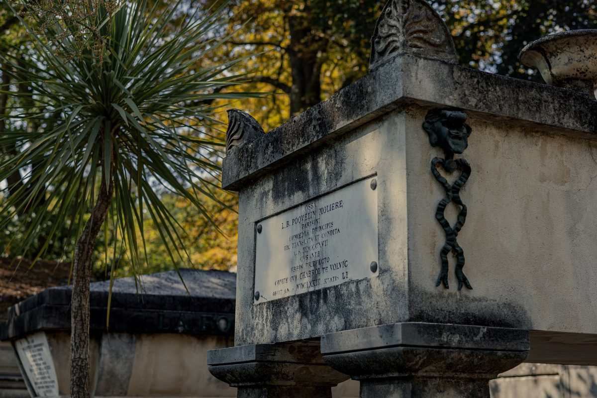 A stone chest tomb with a bronze wreath emblem, a yucca growing behind it