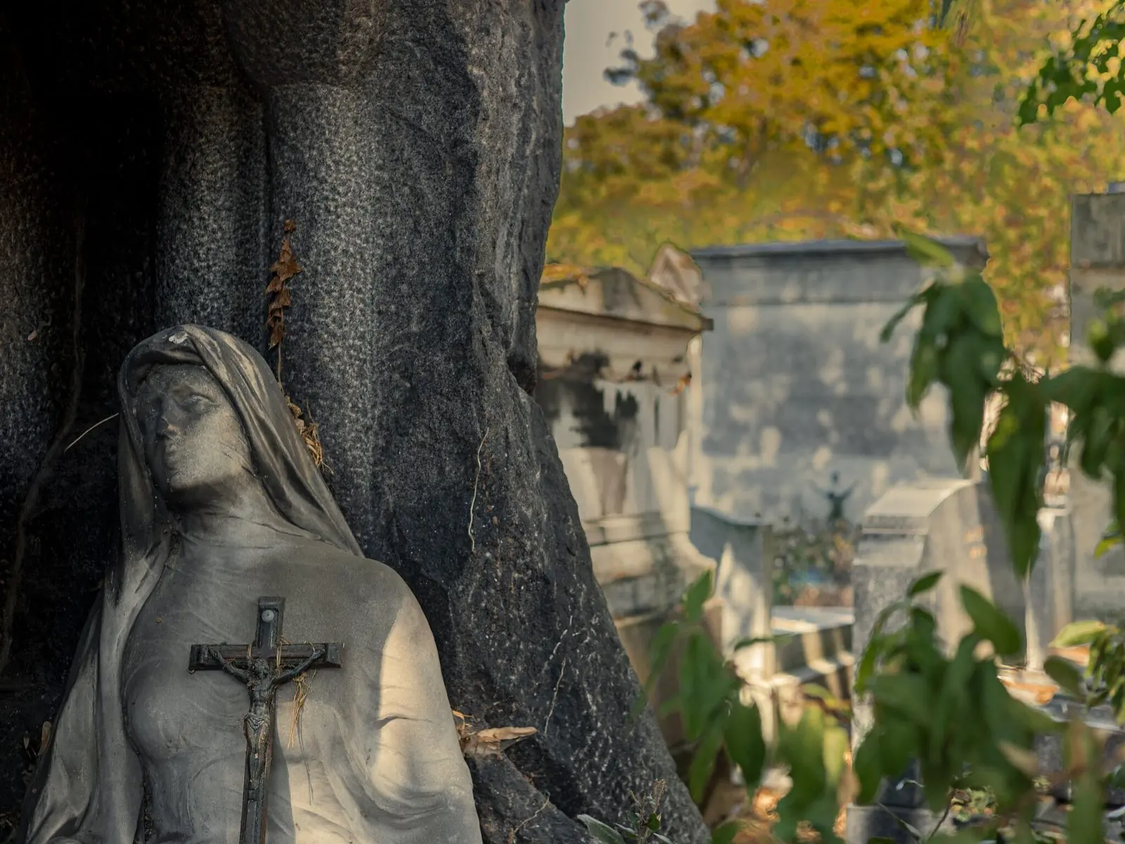A veiled stone mourning figure holding a crucifix against a dark granite monument, sunlit tombs behind
