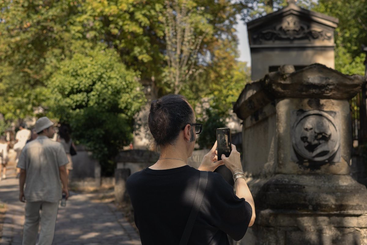 Two visitors on a gravel path, one photographing a tomb carved with a portrait roundel