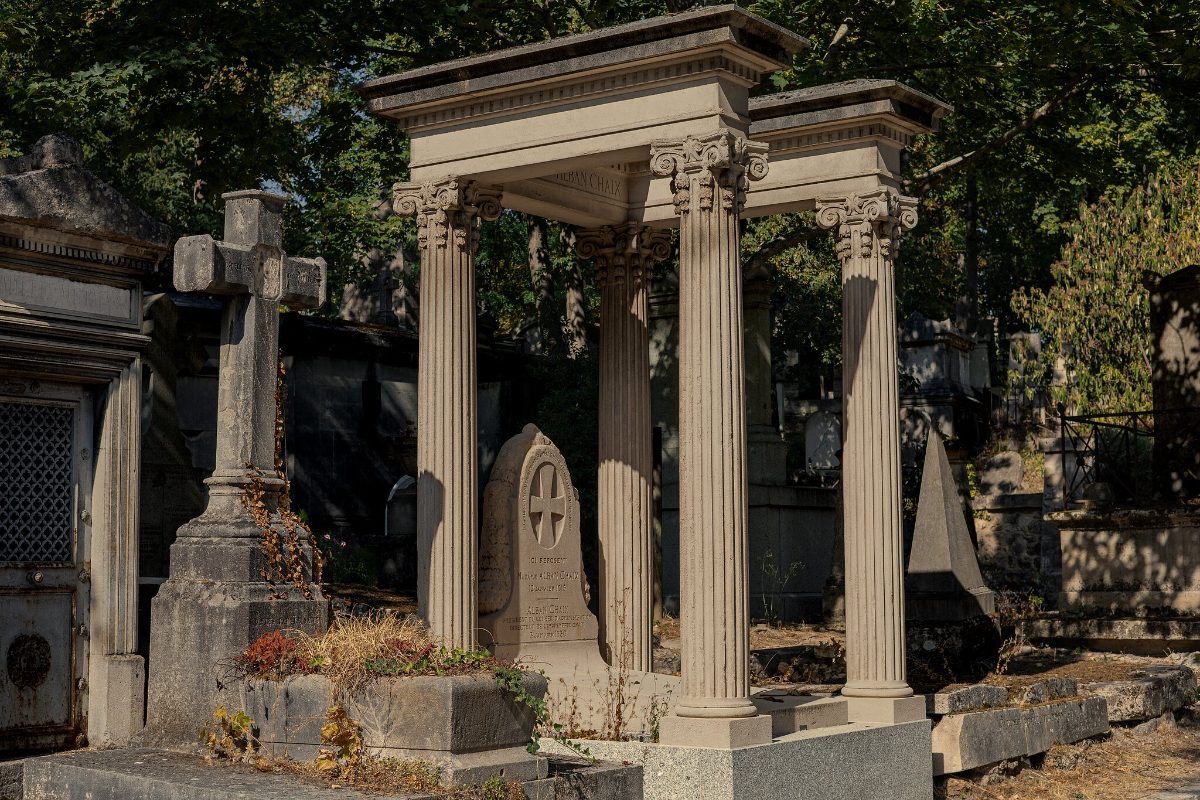 A neoclassical tomb canopy raised on columns, headstones and a stone cross around it